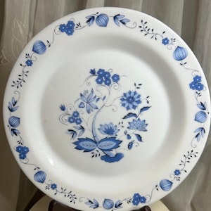 May include: White ceramic plate with a blue floral design. The plate features a central floral arrangement and a border of stylized flowers and leaves. The design is in shades of blue, creating a classic and elegant look. The plate is round.