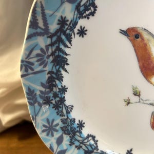 Peut inclure: Une assiette en c&eacute;ramique blanche avec une illustration d&eacute;taill&eacute;e d'un oiseau perch&eacute; sur une branche. Le bord de l'assiette pr&eacute;sente un motif floral bleu et blanc avec des accents bleu fonc&eacute;. L'oiseau a des plumes orange et marron.