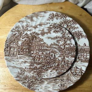 Puede incluir: Dos platos redondos de cerámica con bordes festoneados que presentan un patrón de transferencia en marrón y blanco. El diseño representa una escena rural con un carruaje de caballos, casas de entramado de madera, árboles y un arroyo.