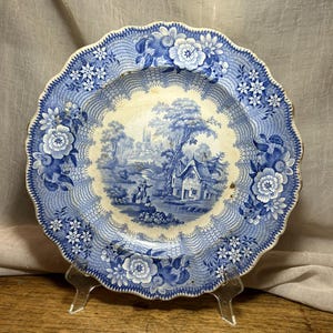 Puede incluir: Un plato de cerámica redondo con borde festoneado, que presenta un patrón de transferencia en azul y blanco. El centro muestra un paisaje pastoral con una cabaña y figuras, mientras que el borde está decorado con motivos florales y de celosía.