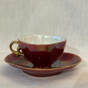 Könnte beinhalten: Ein burgunderfarbenes Teetassen- und Untertassen-Set mit goldenem Griff und Rand. Die Tasse und Untertasse haben eine schillernde Oberfläche, die mehrere Farben reflektiert. Das Set ist wahrscheinlich aus Keramik oder Porzellan.