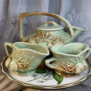 May include: A vintage tea set in green and brown tones, comprising a teapot and two cream jugs. The teapot has a distinctive handle and leaf-like patterns. The set is presented on a decorative tray with floral designs.