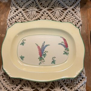 May include: A rectangular cream-coloured ceramic serving platter with scalloped edges and a thin green rim. The centre features a floral pattern of pink and blue delphinium-style flowers with green leaves. A yellow measuring tape is visible.