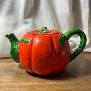 Könnte beinhalten: Eine Keramik-Teekanne in Form eines leuchtend orangefarbenen Kürbisses mit glänzender Oberfläche. Die Teekanne hat einen grünen, strukturierten Griff und Ausguss, die wie Blattreben gestaltet sind, sowie einen passenden grünen Stiel als Deckelgriff.
