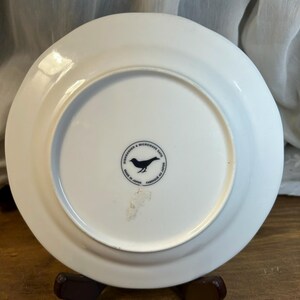 Peut inclure: Une assiette en c&eacute;ramique blanche avec un bord l&eacute;g&egrave;rement sur&eacute;lev&eacute;. Le dessous pr&eacute;sente un logo d'oiseau noir dans un cercle, avec du texte indiquant qu'elle va au lave-vaisselle et au micro-ondes, et fabriqu&eacute;e au Japon.