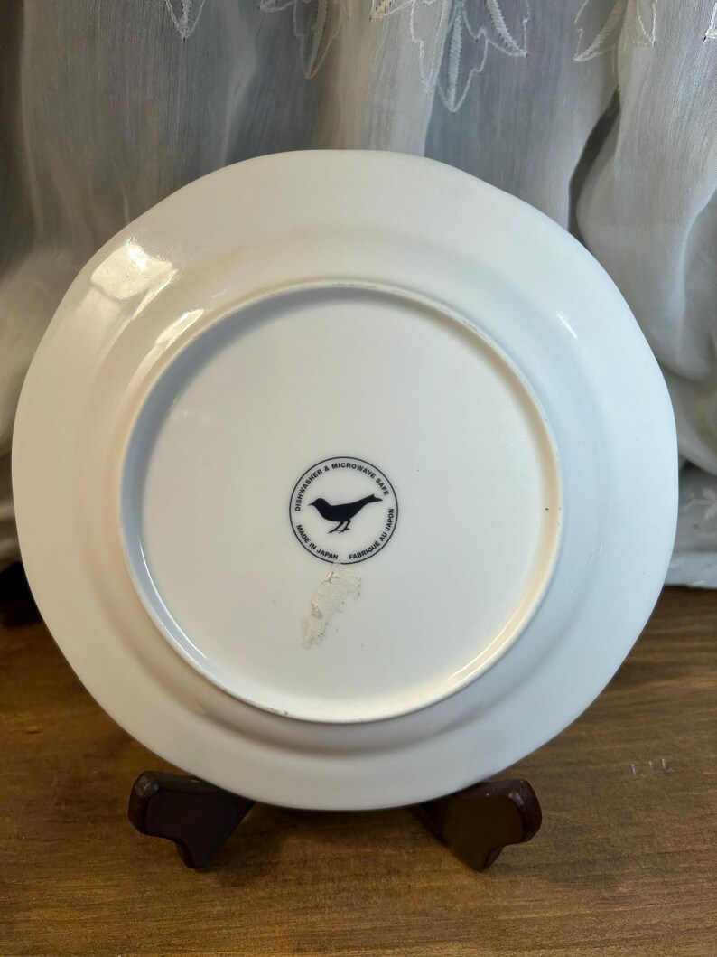 Peut inclure: Une assiette en c&eacute;ramique blanche avec un bord l&eacute;g&egrave;rement sur&eacute;lev&eacute;. Le dessous pr&eacute;sente un logo d'oiseau noir dans un cercle, avec du texte indiquant qu'elle va au lave-vaisselle et au micro-ondes, et fabriqu&eacute;e au Japon.