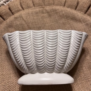 May include: A white ceramic bowl featuring a repeating vertical scalloped or shell-like texture across its exterior surface. The bowl has a wide, flared rim and sits on a solid, smooth, oval-shaped base, presenting a consistent matte finish.