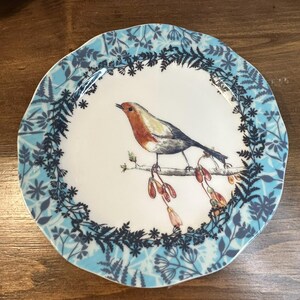 Peut inclure: Assiette ronde en c&eacute;ramique blanche avec une illustration de rouge-gorge sur une branche. L'oiseau a une poitrine rouge et des ailes grises. Le bord de l'assiette pr&eacute;sente un motif floral et de feuilles bleu. Diam&egrave;tre d'environ 20 cm.