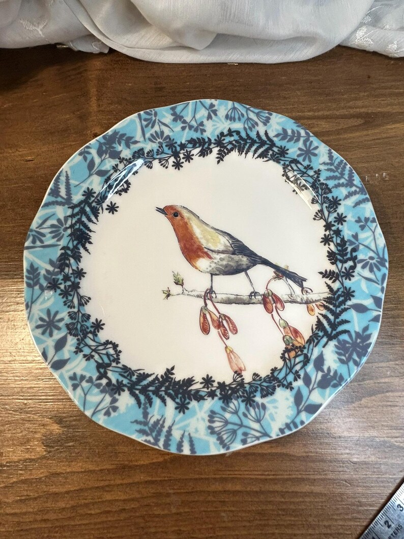 Peut inclure: Assiette ronde en c&eacute;ramique blanche avec une illustration de rouge-gorge sur une branche. L'oiseau a une poitrine rouge et des ailes grises. Le bord de l'assiette pr&eacute;sente un motif floral et de feuilles bleu. Diam&egrave;tre d'environ 20 cm.