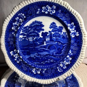 May include: A round ceramic plate with a scalloped edge featuring a blue and white transferware pattern. The central design depicts a landscape with a stone bridge, a castle, and trees. The border is decorated with floral motifs and geometric patterns.