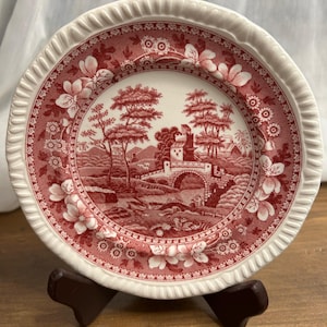 May include: A round plate with a white rim and a red and white landscape design. The plate features a detailed illustration of a bridge, trees, and a building. The plate is displayed on a brown stand.