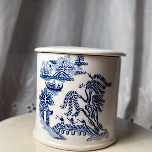 Op de afbeelding: Een witte keramische pot met deksel, met een blauw-wit Willow-patroon. Het patroon toont een landschap met een brug, bomen en figuren. De pot is cilindrisch met een licht uitlopende basis, en de deksel sluit vlak af.