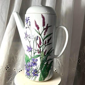 May include: White ceramic coffee pot with a lid, decorated with a floral pattern of purple and pink flowers and green leaves. The pot has a handle and a spout. It is placed on a small, round, white stand.
