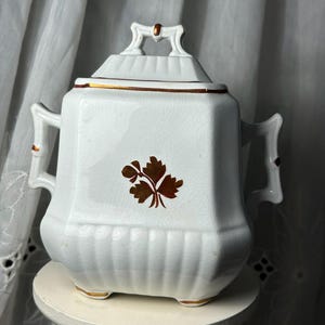 May include: White ceramic sugar bowl with a lid, gold trim, and a brown leaf design. The sugar bowl has a square shape with rounded edges, two side handles, and sits on a small, round base.