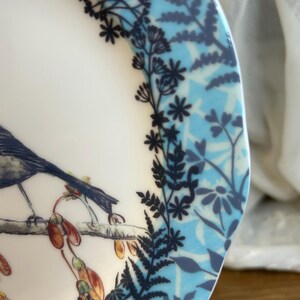 Peut inclure: Une assiette en c&eacute;ramique blanche avec une illustration d&eacute;taill&eacute;e d'un oiseau perch&eacute; sur une branche avec des gousses de graines. Le bord de l'assiette pr&eacute;sente un motif floral bleu et des accents botaniques bleu fonc&eacute;.