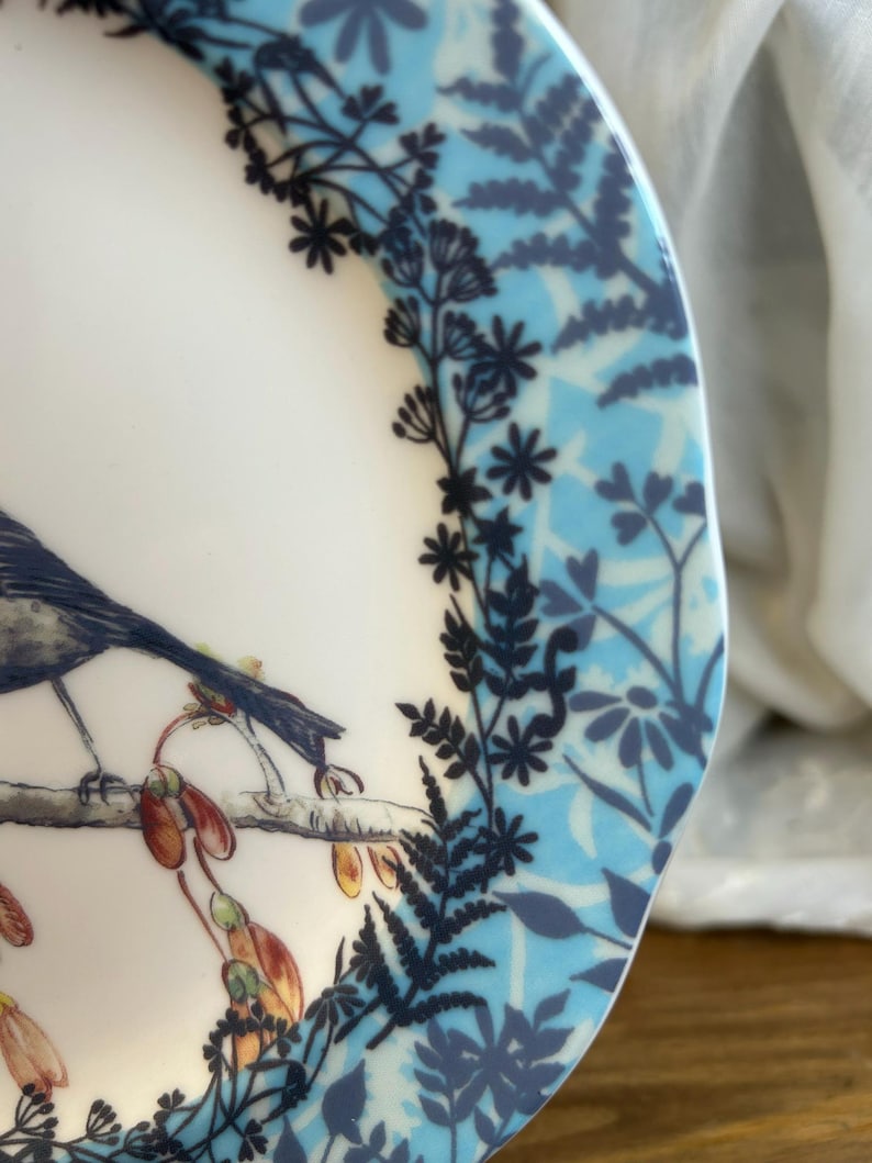 Peut inclure: Une assiette en c&eacute;ramique blanche avec une illustration d&eacute;taill&eacute;e d'un oiseau perch&eacute; sur une branche avec des gousses de graines. Le bord de l'assiette pr&eacute;sente un motif floral bleu et des accents botaniques bleu fonc&eacute;.