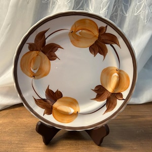 May include: White ceramic plate with a brown rim and scalloped edge. The plate features a hand-painted design of four peaches in shades of orange and brown, with brown leaves. The plate is displayed on a brown wooden stand.