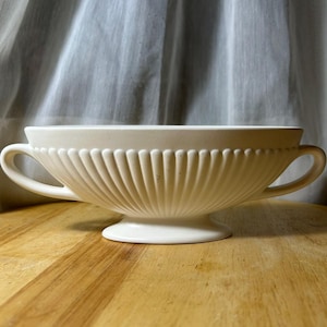 May include: A matte off-white ceramic bowl featuring a wide, shallow basin with vertical fluted ribbing along the exterior. The bowl is supported by a pedestal base and includes two curved handles extending from opposite sides of the rim.