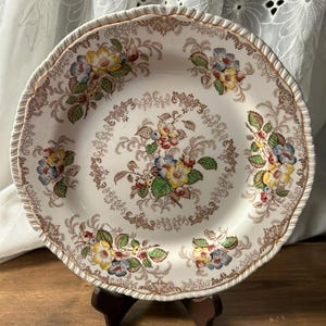 May include: A vintage ceramic plate with a scalloped edge and floral design. The plate features a cream-colored background with brown, yellow, blue, and green floral patterns. The rim has a gold trim. The plate is displayed on a wooden stand.