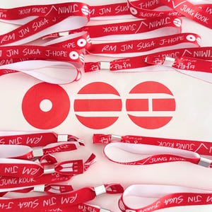Peut inclure: Une collection de bracelets en tissu rouge avec du texte imprimé en blanc incluant les noms RM, JIN, SUGA, J-HOPE, JIMIN et JUNG KOOK, ainsi que le logo BTS. Trois icônes circulaires rouges avec des découpes géométriques blanches sont au centre.