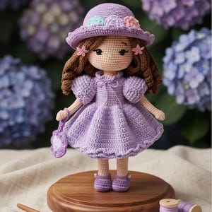 Può includere: Bambola all'uncinetto fatta a mano con un vestito e un cappello color lavanda, adornata con fiori. La bambola ha i capelli ricci castani e porta una piccola borsetta. È su una base di legno, con un uncinetto e rocchetti di filo nelle vicinanze.