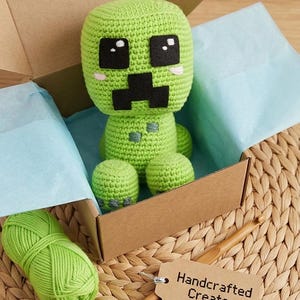 Pode incluir: Um brinquedo Creeper verde de crochê feito à mão, inspirado no Minecraft, apresentado em uma caixa de papelão com papel de seda azul. Uma bola de lã verde, uma agulha de crochê e uma etiqueta com "Handcrafted Creator" também são visíveis.