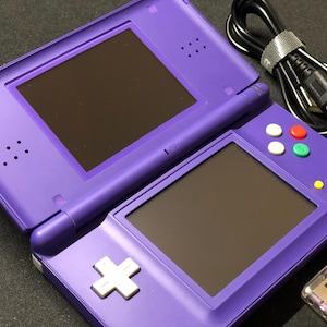 Op de afbeelding: Een paarse Nintendo DS handheld gameconsole, open om twee schermen weer te geven. De console heeft een D-pad, gekleurde knoppen en een stylus. Een zwarte oplaadkabel en een paarse USB-drive zijn ook zichtbaar.