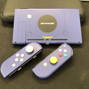 Könnte beinhalten: Eine violette SwitchHouse Nintendo Switch-Konsole mit passenden Controllern. Die Konsole hat einen schwarzen Kreis mit dem Text "SWITCHHOUSE" und einen gelben Akzent. Die Controller haben weiße, rote, grüne und gelbe Knöpfe.