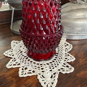 May include: A deep red glass candle holder featuring a prominent diamond-point pattern across its exterior surface. The vessel has a cylindrical shape with a scalloped base and sits atop a white, star-shaped crocheted lace doily with intricate patterns.