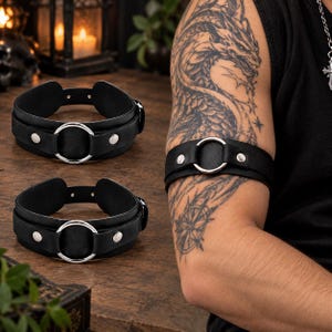 May include: A black leather arm band featuring a central silver-toned metal ring and two silver-toned metal rivets on either side. The band has an adjustable strap design with multiple holes for sizing and a secure buckle closure mechanism.