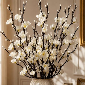 May include: A decorative arrangement of artificial white magnolia flowers with yellow centers, featuring numerous small buds on dark brown, slender, branching stems. The floral display is contained within a textured, light-colored ceramic vase.