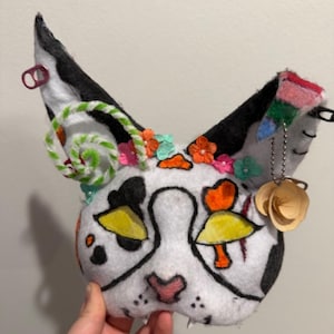 May include: A handmade cat mask with a white base, black ears, and colorful accents. The mask features black spots, yellow and orange details, and is adorned with flowers and a spiral candy. A unique, whimsical costume accessory.