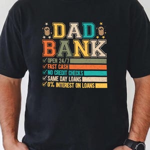 May include: Black t-shirt with a colorful graphic that reads "DAD BANK." The design includes text that says "OPEN 24/7," "FAST CASH," "NO CREDIT CHECKS," "SAME DAY LOANS," and "0% INTEREST ON LOANS." The shirt is a Comfort Colors brand.