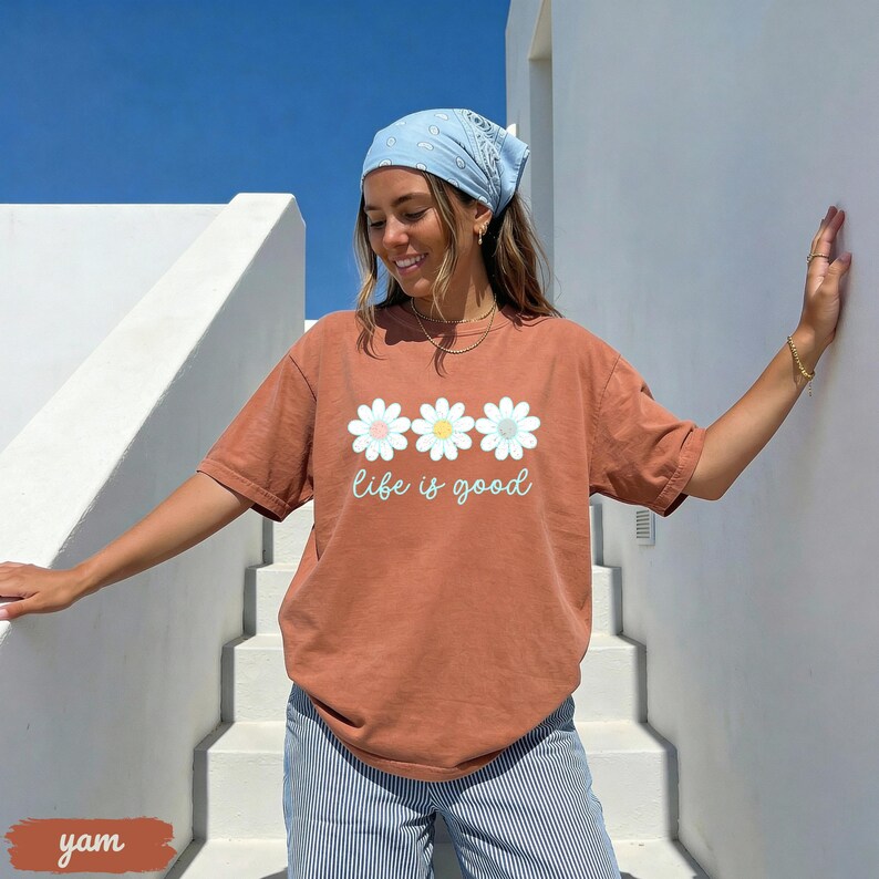 Puede incluir: Una camiseta de color &oacute;xido con la frase "life is good" y tres gr&aacute;ficos de margaritas. Las margaritas son blancas, amarillas y celeste. La persona de la imagen lleva un pa&ntilde;uelo azul claro y pantalones a rayas azules y blancas.
