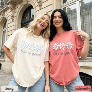 Puede incluir: Dos mujeres con camisetas de color marfil y sand&iacute;a con un gr&aacute;fico de margarita y el texto "life is good". Las camisas se combinan con jeans azules. La camisa marfil tiene texto azul claro y la camisa sand&iacute;a tiene texto rosa oscuro.