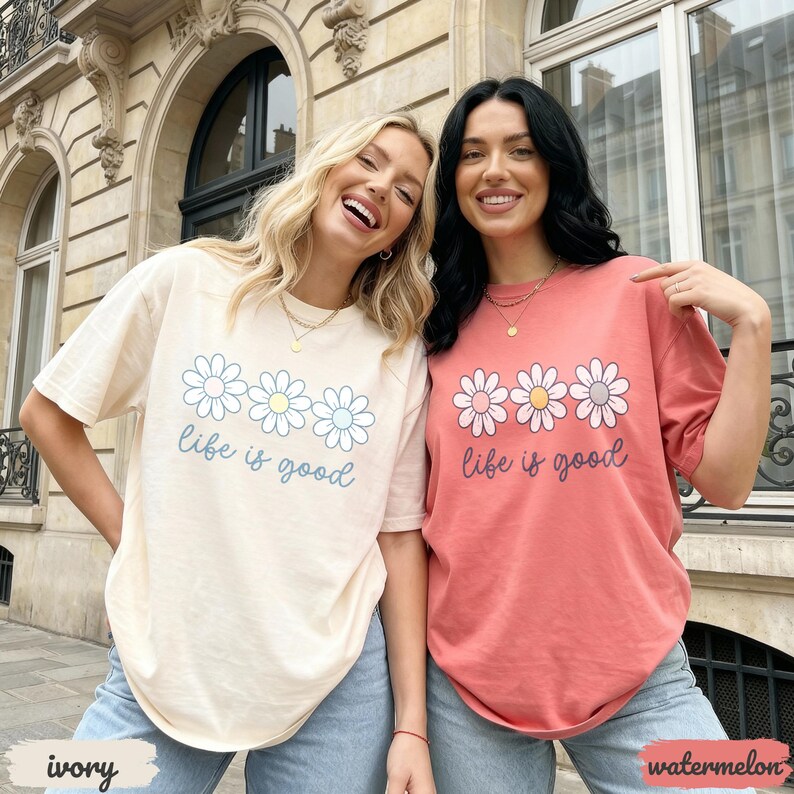 Puede incluir: Dos mujeres con camisetas de color marfil y sand&iacute;a con un gr&aacute;fico de margarita y el texto "life is good". Las camisas se combinan con jeans azules. La camisa marfil tiene texto azul claro y la camisa sand&iacute;a tiene texto rosa oscuro.