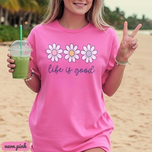Puede incluir: Camiseta rosa ne&oacute;n con un dise&ntilde;o de margaritas y la frase "life is good". Las margaritas tienen p&eacute;talos blancos y centros rosas, amarillos y morados. La camiseta es de un color vibrante y tiene un corte holgado.