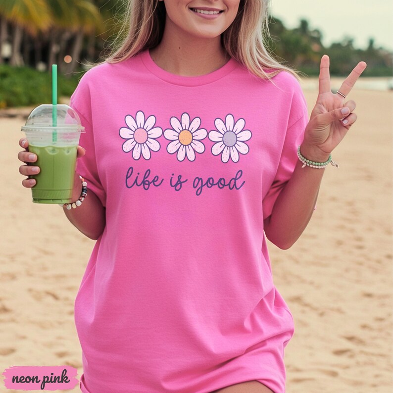 Puede incluir: Camiseta rosa ne&oacute;n con un dise&ntilde;o de margaritas y la frase "life is good". Las margaritas tienen p&eacute;talos blancos y centros rosas, amarillos y morados. La camiseta es de un color vibrante y tiene un corte holgado.