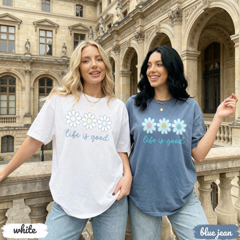 Puede incluir: Dos mujeres con camisetas blancas y azul vaquero. La camiseta blanca tiene un dise&ntilde;o de margarita con el texto "life is good" en azul claro. La camiseta azul tiene un dise&ntilde;o y texto similares. Ambas son de manga corta.
