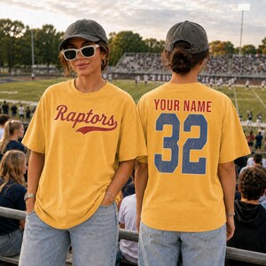 A woman wearing a yellow t-shirt with the word "Raptors" and the number 32, standing in a stadium with a crowd in the background.