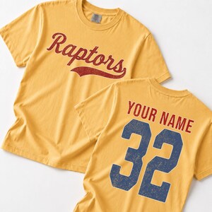 A yellow t-shirt with the word "Raptors" printed in red cursive text, and the number "32" printed in blue.