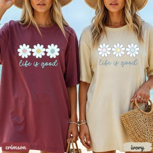 Puede incluir: Dos camisetas de gran tama&ntilde;o, una carmes&iacute; y otra marfil, cada una con un dise&ntilde;o de margarita y la frase "life is good". El dise&ntilde;o de la margarita incluye colores pastel. Las camisetas tienen mangas cortas y un corte holgado.