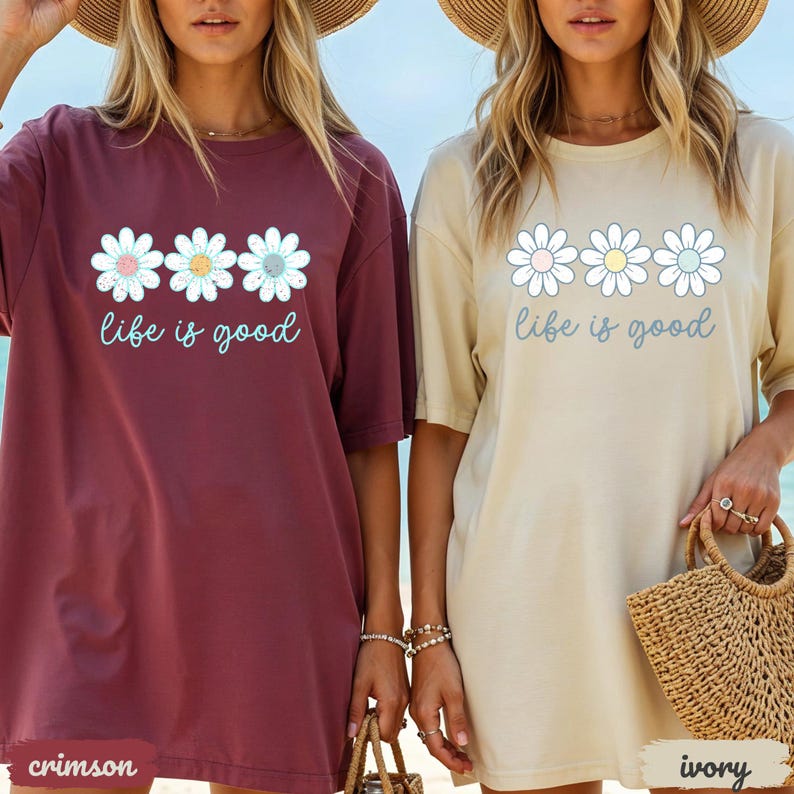 Puede incluir: Dos camisetas de gran tama&ntilde;o, una carmes&iacute; y otra marfil, cada una con un dise&ntilde;o de margarita y la frase "life is good". El dise&ntilde;o de la margarita incluye colores pastel. Las camisetas tienen mangas cortas y un corte holgado.