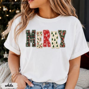 May include: A white short-sleeve t-shirt featuring the word MERRY in large block letters across the chest. Each letter has a unique holiday pattern: green plaid with stars, holly berries, red with white snowflakes, pine trees, and candy canes.