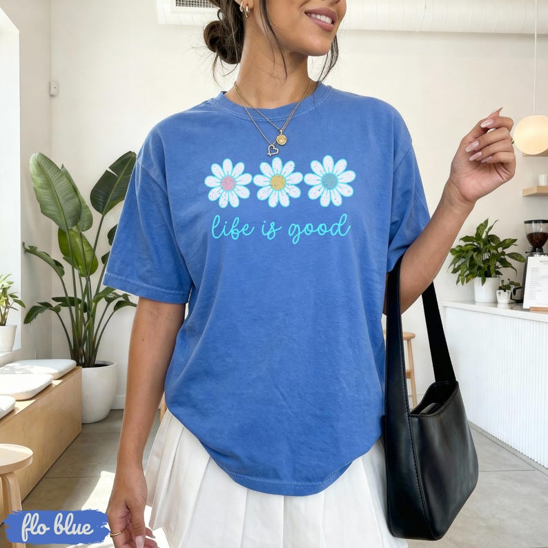 Puede incluir: Camiseta azul con la frase "life is good" en fuente cursiva. La camiseta presenta tres gr&aacute;ficos de flores de margarita con centros rosas, amarillos y azules. La camiseta es de color azul liso.