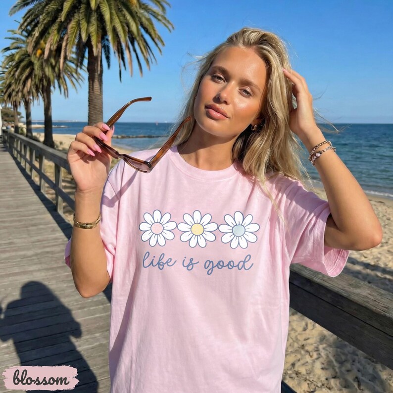 Puede incluir: Camiseta rosa claro con un dise&ntilde;o de margaritas y la frase "life is good". La camiseta se usa al aire libre en un d&iacute;a soleado, con una playa de fondo.