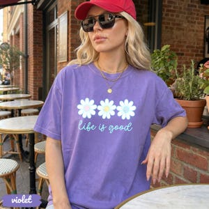 Puede incluir: Una camiseta violeta con un dise&ntilde;o de margarita y el texto "life is good". El dise&ntilde;o de la margarita tiene p&eacute;talos blancos y centros azul claro y amarillo. La persona de la imagen lleva una gorra de b&eacute;isbol roja y gafas de sol.