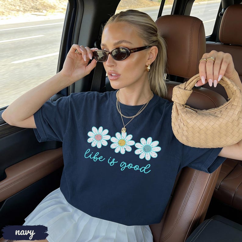 Puede incluir: Camiseta azul marino con tres flores de margarita y la frase "life is good" en turquesa. La persona lleva gafas de sol y joyas doradas, y sostiene un bolso beige. La camisa se combina con una falda blanca.
