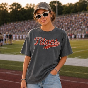 A young woman wearing a gray t-shirt with the word "Titans" and sunglasses stands on a sports field.
