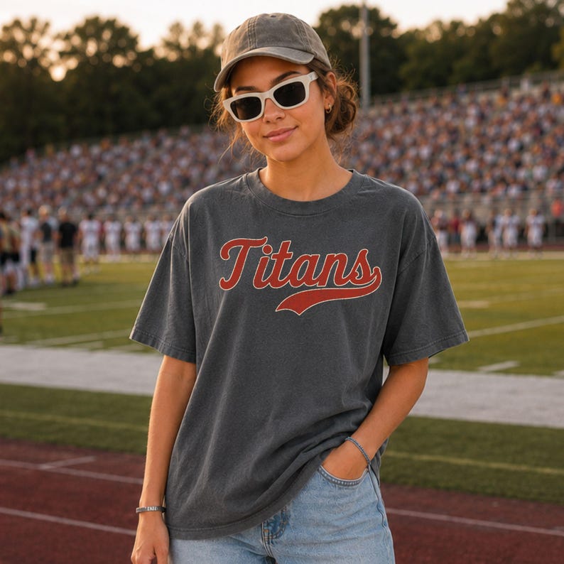 A young woman wearing a gray t-shirt with the word "Titans" and sunglasses stands on a sports field.
